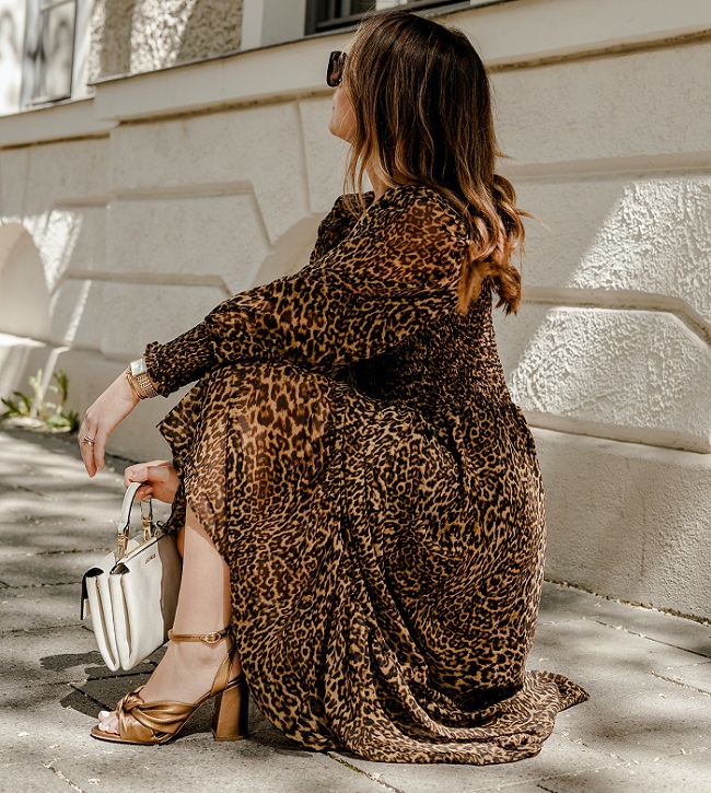 picture of a woman standing on a sidewalk wearing dress with print, brown dress sandals, holding white bag